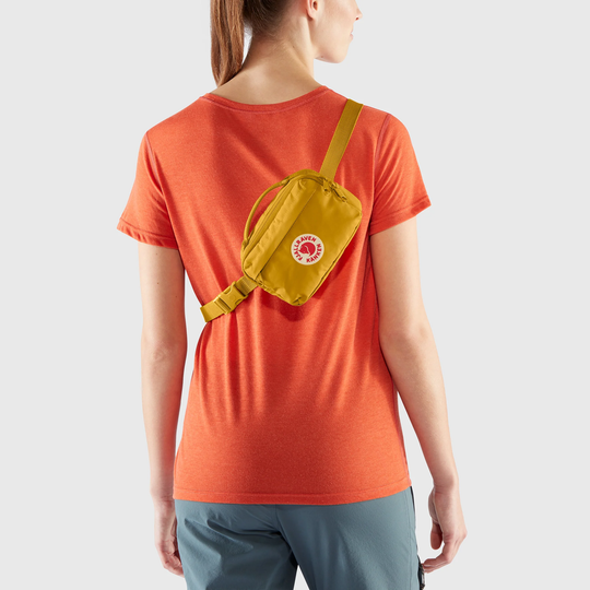 Kånken Hip Pack in mustard color worn around waist on an orange shirt, featuring durable Vinylon F fabric and multiple pockets.