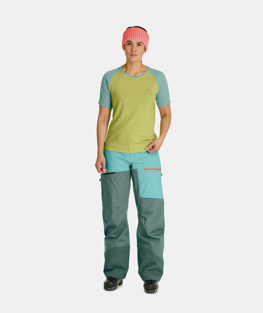 Woman wearing 150 Cool Ballpen TS, green Merino T-shirt and outdoor pants, standing with a pink beanie.
